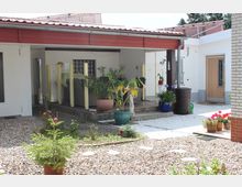 Ein Innenhof mit einem überdachten Sitzbereich, umgeben von weißen Wänden und großen Blumenkübeln mit verschiedenen Pflanzen. Der Boden ist teilweise mit Kieselsteinen bedeckt, und es gibt eine Holztür auf der rechten Seite sowie ein Regenfass und bunte Blumen in Töpfen.