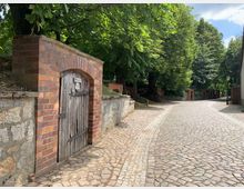 Gepflasterte Straße mit geschwungenem Verlauf, gesäumt von alten Bäumen und einer niedrigen Mauer aus Stein und Ziegeln. Im Vordergrund ein Holztor in einem gemauerten Torbogen, während der Weg in einer ruhigen, bewaldeten Umgebung verläuft.