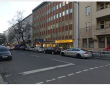 Straßenansicht mit mehreren Wohn- und Geschäftsgebäuden, darunter ein Gebäude mit einer rot-beigen Fassade, in dem sich ein Geschäft namens "Juwelier Tülger" befindet. Auf der Straße parken mehrere Autos, und links ist ein Eingang zur U-Bahn-Station "Bismarckstraße" erkennbar.