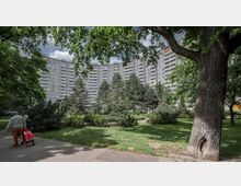 Gebogene Wohnanlage mit mehreren Etagen im Hintergrund, umgeben von Bäumen und dichter Grünfläche; im Vordergrund ein Gehweg mit zwei Personen, von denen eine einen roten Einkaufstrolley zieht.