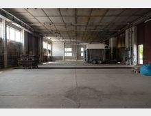 Eine leere, industrielle Werkhalle mit hohen Decken und einfachen Betonböden. Im Hintergrund steht ein Anhänger, umgeben von gestapelten Paletten und industriellen Geräten, während große Fenster auf der linken Seite und ein offenes Tor auf der rechten Seite Tageslicht hereinlassen.