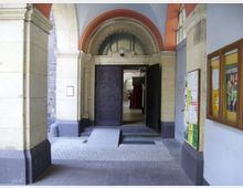 Eingangsbereich eines historischen Gebäudes mit Rundbogen und verzierten Doppeltüren. Neben dem Eingang befinden sich Informationstafeln und ein Aushangkasten, der Innenraum ist teilweise sichtbar und zeigt hohe Decken mit Fenstern.
