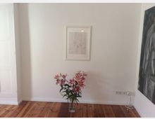 Ein minimalistischer Innenraum mit einer weißen Wand, an der ein gerahmtes Kunstwerk hängt. Auf dem Holzboden steht eine Vase mit rosa Lilien, und an der Wand befinden sich Steckdosen und ein loses Kabel.