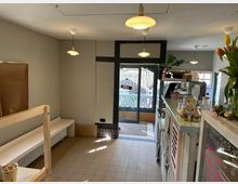 Kleines Café mit heller Einrichtung, bestehend aus einer weißen Theke mit Dekorationen und Snacks, einer Sitzbank entlang der Wand sowie einem Glaseingang mit Blick auf eine kleine Terrasse. Mehrere Hängelampen beleuchten den gekachelten Boden und die gemütliche Atmosphäre.