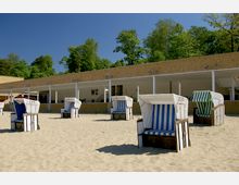Ein Sandstrand mit mehreren Strandkörben, die blau-weiß oder grün-weiß gestreift sind, vor einem niedrigen, gelben Gebäude mit überdachtem Bereich und der Aufschrift "WC". Im Hintergrund sind Bäume und ein klarer Himmel zu sehen.