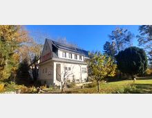 Zweistöckiges Wohnhaus mit hohem Satteldach und Solarpaneelen, umgeben von einem großen Garten mit Rasenfläche und Bäumen. Im Vordergrund sind ein kleiner Baum, Beete und ein schmaler Weg entlang der Hauswand zu sehen.