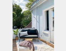 Gemütliche Terrasse mit Holzboden und einer modernen Sitzbank mit dunklen Kissen, ergänzt durch einen kleinen Holztisch und eine Pflanze im Korb. Im Hintergrund sind grüne Bäume und Teile eines Gebäudes mit Ziegeldach sichtbar.