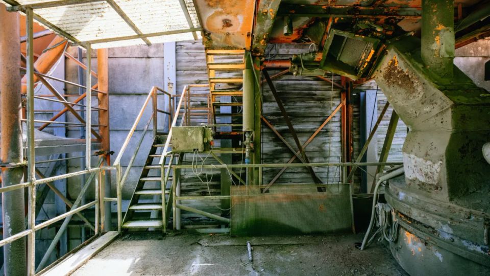 Industrielle Anlage mit rostigen Metalltreppen, Geländern und Rohrleitungen, umgeben von verwitterten Wänden und einer teils durchlässigen Überdachung. Die Szenerie zeigt Gebrauchsspuren wie Rost und abgeblätterte Farbe.