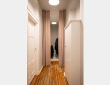 Kleiner Flur mit Holzdielenboden, rechts ein weißer Schrank und links eine weiße Tür. Am Ende des Flurs hängt ein beigefarbener Vorhang, der einen Blick auf einen Raum mit Kleiderständern freigibt.