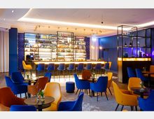 Ein modernes Bar-Interieur mit einer beleuchteten Theke und hohen Regalen voller Flaschen im Hintergrund. Der Raum ist mit blauen, gelben und orangefarbenen Polsterstühlen ausgestattet, und kleine runde Tische stehen auf einem grauen Teppich.