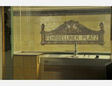 Retro U-Bahn-Station mit gelb gefliester Wand und der Aufschrift „Fehrbelliner Platz“ in einem kunstvoll eingerahmten Schild; im Vordergrund ein kleiner Bedienpult mit Schaltern und Holzverkleidung.