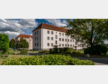 Mehrfamilienhaus mit hellbeiger Fassade und roten Ziegeldächern in einer Wohngegend, umgeben von gepflegtem Grün, Bäumen und einem Gehweg. Im Vordergrund sind Büsche und ein Straßenlaternenmast zu sehen, während der Himmel von Wolken durchzogen ist.