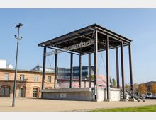 Freiluftbühne mit einer Überdachung aus Stahlträgern, umgeben von einem gepflasterten Platz. Im Hintergrund sind ein Backsteingebäude und moderne Bürogebäude sichtbar, umgeben von Herbstbäumen unter klarem blauem Himmel.