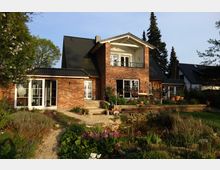 Zweistöckiges Einfamilienhaus aus rotem Backstein mit dunklem Satteldach, großem Balkon und großen Fenstern. Im Vordergrund befinden sich ein gepflegter Garten mit Blumenbeeten, ein kleiner Teich sowie eine Terrasse mit Pflanzen.