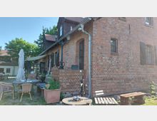 Backsteingebäude mit schwarzen Fensterläden und Satteldach, umgeben von einem gepflasterten Außenbereich mit Tischen, Stühlen und einem Sonnenschirm. Im Vordergrund stehen eine alte Wasserpumpe, Blumenkübel und eine Holzbank.