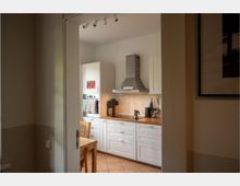 Blick durch eine offene Tür in eine helle Küche mit weißen Schränken, Holzarbeitsplatte und Edelstahl-Dunstabzugshaube über dem Herd. Fliesenboden, kleine Küchenutensilien auf der Arbeitsfläche und ein Holztisch mit Stühlen sind sichtbar.