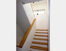 Eine gerade Innentreppe mit hellen Holzstufen und passendem Handlauf. Die Wände sind weiß, und oben befindet sich ein kleines rechteckiges Fenster mit einer Pflanze auf der Fensterbank.