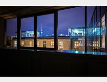 Blick durch ein großes Fenster auf ein beleuchtetes Gebäude mit Fenstern und Flachdach, im Hintergrund der Fernsehturm bei Nacht. Die Szene zeigt eine Mischung aus urbaner Architektur und moderner Beleuchtung.