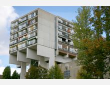 Wohnblock in moderner Architektur mit mehreren Stockwerken, schwebend auf massiven Betonpfeilern gebaut. Die Fassade zeigt zahlreiche Balkone mit Geländern, Satellitenschüsseln und einfarbigen Markisen, umgeben von Bäumen.