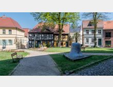 Ein kleiner Platz mit Kopfsteinpflaster, zwei Bänken und einer großen Glocke auf einem Podest. Umgeben von Fachwerk- und Wohnhäusern mit roten Ziegeldächern; im Hintergrund befinden sich Bäume und Grünflächen.