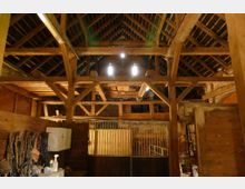 Holzinterieur eines alten Stalls mit sichtbarem Dachgebälk und Satteldachkonstruktion. Im Vordergrund sind Holzboxen mit Gittertüren sowie an der Wand aufgehängtes Reitzubehör und Geräte erkennbar.