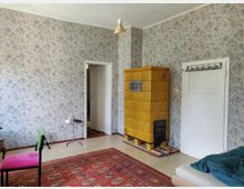 Ein schlicht eingerichtetes Zimmer mit blumengemusterten Tapeten, einer gelben Kachelofenheizung, einer weißen Tür und einem roten Teppich mit orientalischem Muster. Im Vordergrund stehen ein Stuhl mit farbigem Sitzpolster, ein kleiner Beistelltisch und eine Matratze mit blauer Decke.