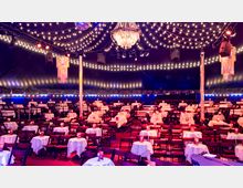 Innenansicht eines festlich dekorierten Veranstaltungszelts mit gedeckten Tischen und Klappstühlen. Eine Kuppeldecke mit Lichterketten und ein großer Kronleuchter bilden die Hauptbeleuchtung, mit zusätzlicher Dekoration wie kleineren Hängeleuchtern.