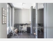 Modernes Arbeitszimmer mit grauen Wänden und dunklem Holzboden. Der Raum ist eingerichtet mit einem schlichten Schreibtisch, einem weißen Designerstuhl, Metallregalen und einem Korb; große Fenster auf der linken Seite lassen viel Tageslicht herein.