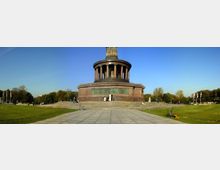 Eine monumentale Säule mit einer runden, von Säulen getragenen Plattform auf einem quadratischen Sockel aus Stein, umgeben von einem großen gepflasterten Platz und Rasenflächen, mit Bäumen im Hintergrund unter klarem blauem Himmel.