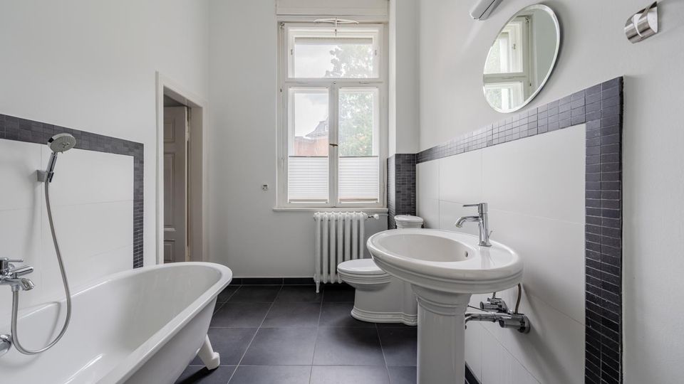 Helles Badezimmer mit freistehender Badewanne, Waschbecken auf Säule und WC, weiß gefliest mit dunkler Fliesenbordüre und dunklem Boden. Ein großes Fenster mit Heizkörper darunter sowie ein runder Wandspiegel und eine Leuchte ergänzen den Raum.