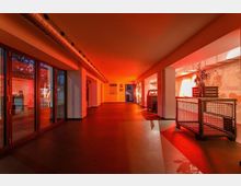 Ein modern gestalteter Innenraum mit roten Lichteffekten, großen Glasfenstern auf der linken Seite und freiliegenden Lüftungsrohren an der Decke. Rechts befinden sich Mauerwerkdetails sowie dekorative Holz- und Metallmöbel.