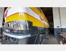 Gelbe Lokomotive mit schwarz-weißen Streifen und großem Logo, abgestellt in einem Gebäude mit weißen Ziegelwänden und Holzdach. Vorderseite der Lok mit glänzenden Metallapplikationen und Scheinwerfern im Fokus.