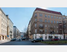 Straßenszene mit mehrstöckigen Altbau-Wohnhäusern, teils mit kleinen Balkonen. Im Erdgeschoss des Eckgebäudes befinden sich Geschäfte, während parkende Autos und einzelne Fußgänger im Vordergrund zu sehen sind.