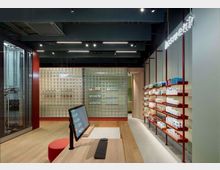 Innenansicht einer modernen Apotheke mit einem hellen, offenen Design. Der Raum ist durch eine Wand aus Glasbausteinen geteilt, Regale mit Produkten verlaufen entlang der rechten Seite, während ein Monitor und ein Schreibtisch im Vordergrund stehen.