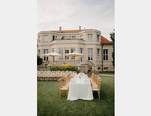 Großer Garten vor einer eleganten Villa mit hellem Putz, vielen hohen Fenstern und rotem Ziegeldach. Auf dem Rasen stehen lange, weiß gedeckte Tafeln mit Holzstühlen, daneben Sonnenschirme, Blumenbeete und eine Steinterrasse mit Treppen.