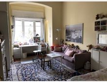 Heller Wohnraum mit beiger Wandfarbe, großer Fensterfront mit weißem Vorhang, einer Arbeitsecke mit Schreibtisch und Stuhl sowie einer Sitzecke mit grauem Sofa, Couchtisch und einem Teppich mit Muster. Regale und Dekorationsgegenstände ergänzen die Einrichtung.