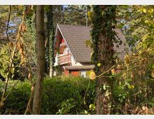 Ein Haus mit Satteldach und rotem Balkon, umgeben von dichtem Wald und üppigem Grün. Die Fassade ist teils von Sonnenlicht beleuchtet, während Bäume und Kletterpflanzen das Gebäude teilweise verdecken.