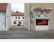 Eine kleine Ladenfassade eines Möbelmarkts mit roten Schriftzügen, die einen Total-Ausverkauf ankündigen. Im Hintergrund ein Innenhof mit einem geparkten Auto vor einem weiteren Gebäudeteil mit weißen Wänden und roten Dachziegeln.