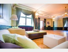 Ein eleganter, heller Innenraum mit hohen Decken, einem Holzboden, cremefarbenen Sofas und einem weißen Couchtisch. Große Fenster mit grünen Vorhängen, verziertes Stuckdesign an der Decke und ein Kronleuchter prägen den Raum, der mit dekorativen Säulen und einer Tür im Hintergrund versehen ist.