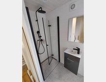 Modernes Badezimmer mit bodengleicher Dusche und Glasabtrennung, weißen Wandfliesen und grauem Fliesenboden. Schwarze Duscharmatur mit Regendusche sowie ein kleiner Waschtisch mit Spiegel und schwarzem Wasserhahn sind zu sehen.