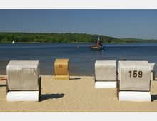 Strand mit mehreren Strandkörben auf hellem Sand, dahinter ein ruhiger See mit einem Steg und einer Wasserrutsche sowie einer grünen Uferlandschaft im Hintergrund. Ein Segelboot ist auf dem Wasser zu sehen, der Himmel ist klar und blau.