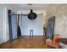 Fotostudio in einem Raum mit verputzten Wänden und Fischgrät-Parkettboden. Ein Beleuchtungsstativ mit Softbox, eine graue Hintergrundleinwand, ein Hocker und Requisiten wie bemalte Platten und ein rosafarbener Sessel sind sichtbar.