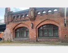 Backsteinbau im historischen Stil mit Spitzbogenfenstern und Gittern, gekennzeichnet durch die Aufschrift „Friedrichshagen“. Links ist ein kleiner Turm zu sehen, rechts eine angebaute Laterne, im Hintergrund verläuft eine Brücke.