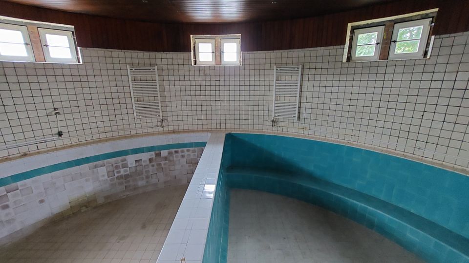 Innenansicht eines leerstehenden, ovalen Pools mit türkisfarbenen Fliesen und einer umlaufenden weißen Fliesenumrandung. Die Wände sind mit weißen Kacheln verkleidet, es gibt mehrere kleine Fenster und zwei Heizkörper an den Wänden.