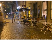 Nächtliche Szene einer schmalen Kopfsteinpflasterstraße mit Außengastronomie: Klapptische und Stühle stehen entlang einer Gebäudefassade unter großen Sonnenschirmen, beleuchtet von Laternen und warmem Innenlicht aus Fenstern. Pflanzkübel und Dekoelemente säumen den Rand, im Hintergrund setzt sich die Straße mit weiteren Leuchten und Sitzbereichen fort.