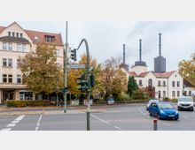 Straßenecke mit historischen Wohngebäuden, Herbstbäumen und einem Straßenschild mit der Aufschrift "Ostpreußendamm". Im Hintergrund sind moderne Industriekamine sichtbar, während im Vordergrund Autos an einer Ampel warten.