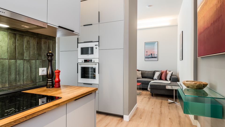Offener Wohnbereich mit moderner Küche: helle, grifflose Schränke, Holzarbeitsplatte und grüne Fliesenrückwand mit eingebautem Ofen und Mikrowelle. Dahinter schließt ein kleines Wohnzimmer mit grauem Ecksofa an; rechts ein Glas-Wandregal und gerahmte Bilder an weißen Wänden über hellem Holzboden.
