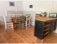 Heller Essbereich mit Holzdielenboden, weiß vertäfelten Wänden und einem rechteckigen Holztisch, umgeben von gemischten Holzstühlen. Rechts steht ein Küchenblock mit Holzarbeitsplatte und Schubladen, daneben ein kleines Regal; an der Wand hängen zwei Bilder und eine Pendelleuchte.