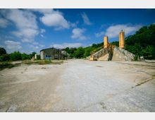 Weitläufiges Gelände mit Betonboden, einer verlassenen Industrieanlage mit rostfarbenen Silotürmen rechts und einem kleinen, verfallenen Gebäude links, umgeben von vereinzelter Vegetation und Wald im Hintergrund unter blauem Himmel.