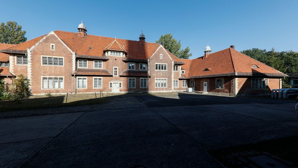 Großer Backsteinbau mit roten Ziegeldächern und mehreren kleinen Dachaufsätzen, frontseitig mit vielen rechteckigen Fenstern und einem mittigen Eingangsbereich. Davor liegt ein gepflasterter Hof, umgeben von Bäumen unter klarem blauem Himmel.